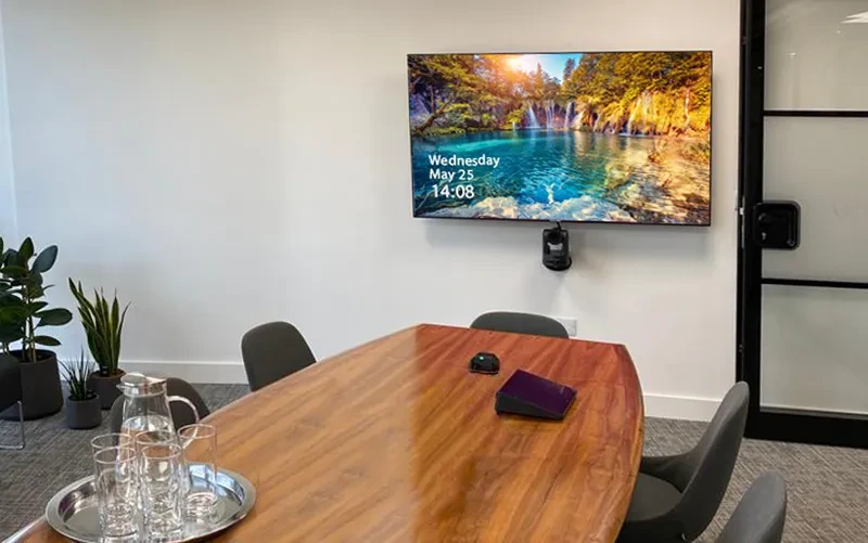 A conference room with a wall-mounted display and video conferencing camera, positioned above a boardroom table.