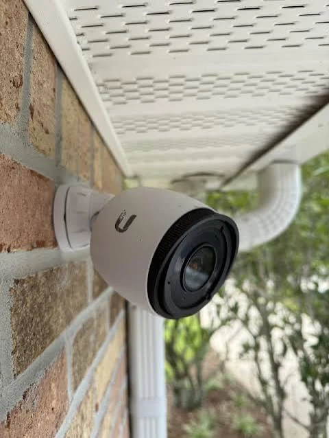 An outdoor security camera mounted under the eave of a brick house, angled to cover the walkway and yard.